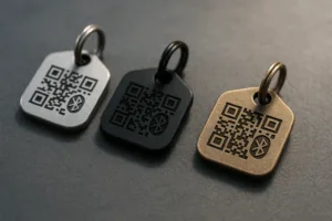 A selection of dog tag trackers on display, featuring brushed nickel, matte black, and antique brass plating, with engraved QR codes and Bluetooth symbols.