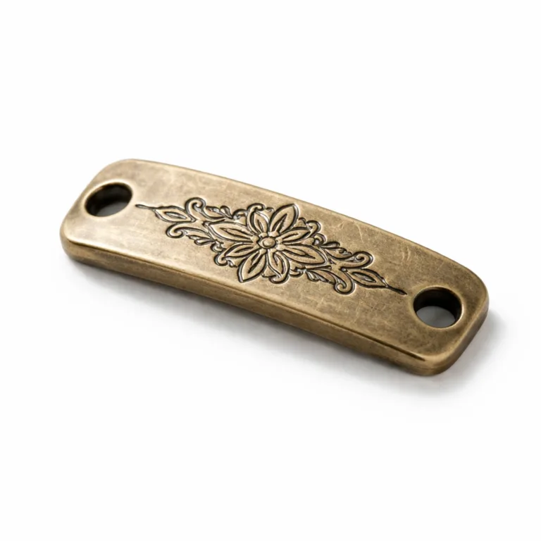 An artisan bracelet tag made from antique brass with a tactile sandblasted matte texture and thicker profile. The front displays a finely etched pattern while the double sewing holes enable simple attachment to fabric or leather bands. Presented in high definition for e-commerce on a white studio backdrop.