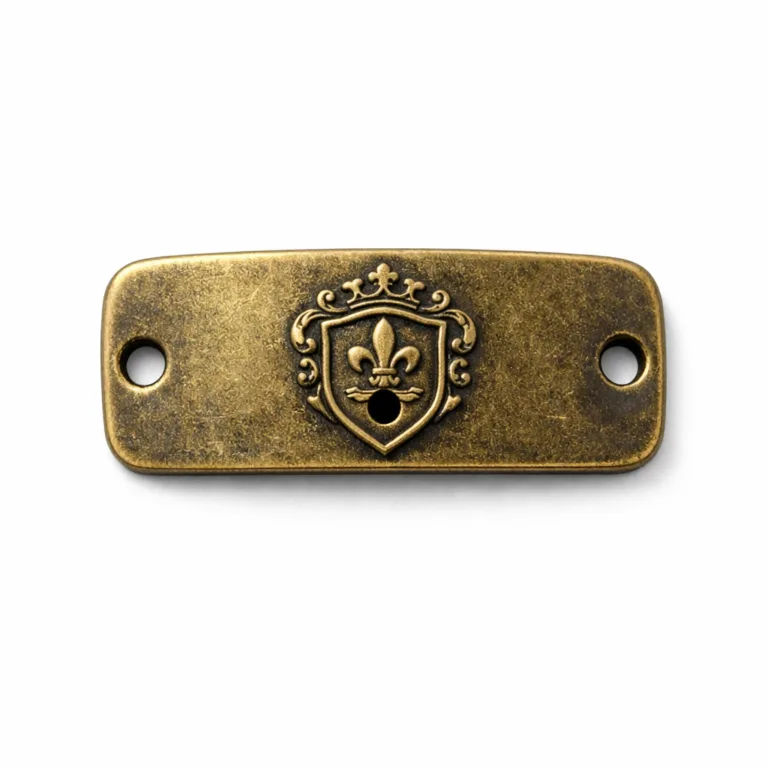 A highly realistic shot depicting an antique brass bracelet tag plate with smooth rounded corners, an embossed front emblem, and a single central sewing hole. The classic finish and design highlight the tag's vintage appearance for stylish jewelry.
