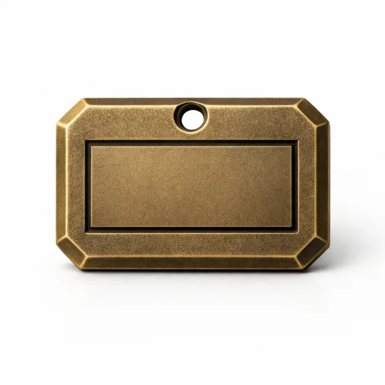 Die-cast bag tag emblem featuring an antique brass finish, sandblasted matte texture, chamfered edge detailing, and an etched blank logo panel with a single hole for easy chain attachment.
