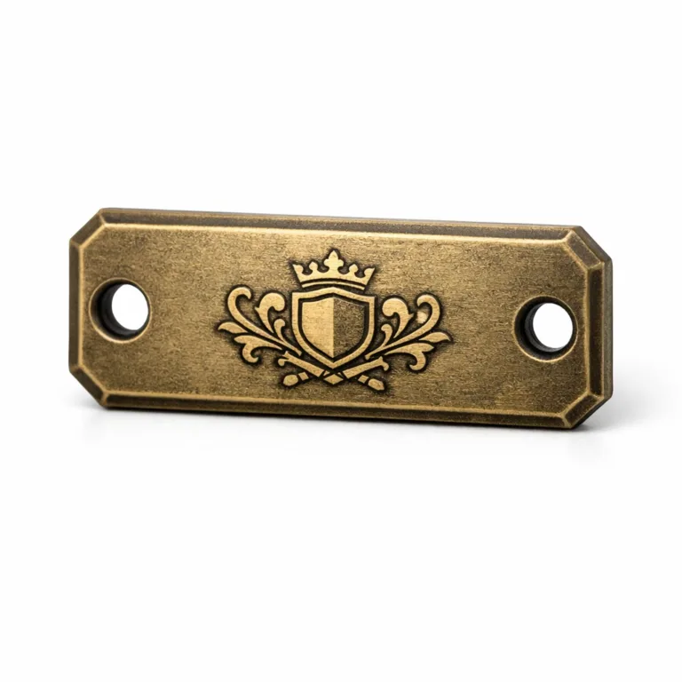 A matte nickel garment tag styled with an antique brass finish, thick chamfered profile, double sewing holes, and an etched emblem design for a vintage and premium aesthetic.