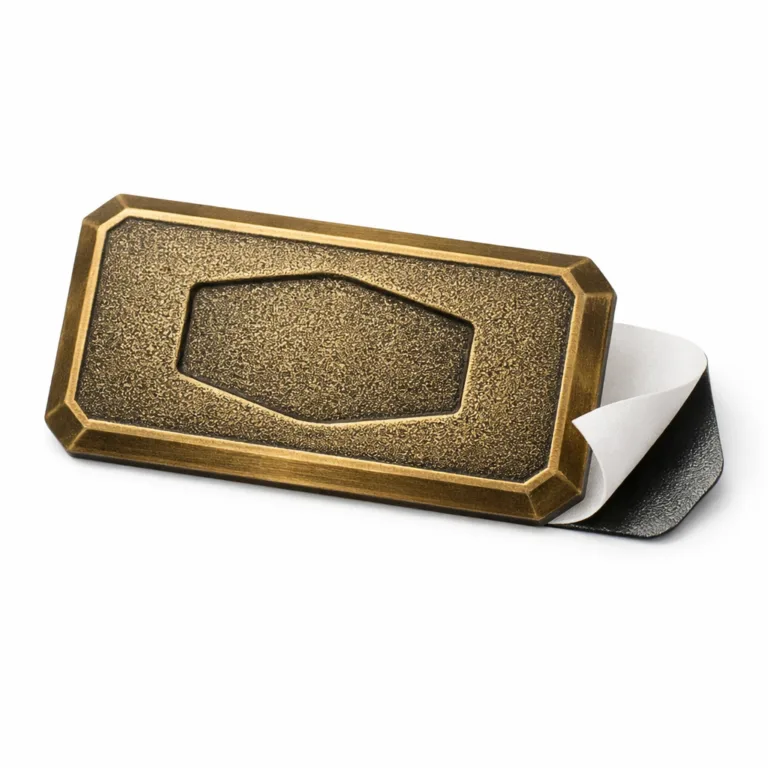 This logo tag is crafted from metal with an antique brass finish, featuring chamfered edges, a debossed emblem design, and a visible adhesive backing for easy attachment to various surfaces.