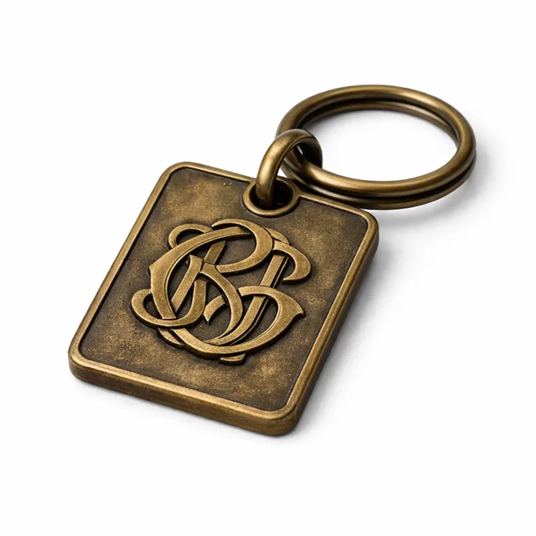 One monogram bag tag in an antique brass finish, showcasing an embossed logo with beveled edges and a matching brass split ring. The worn patina gives a classic vintage look with dimensional branding.