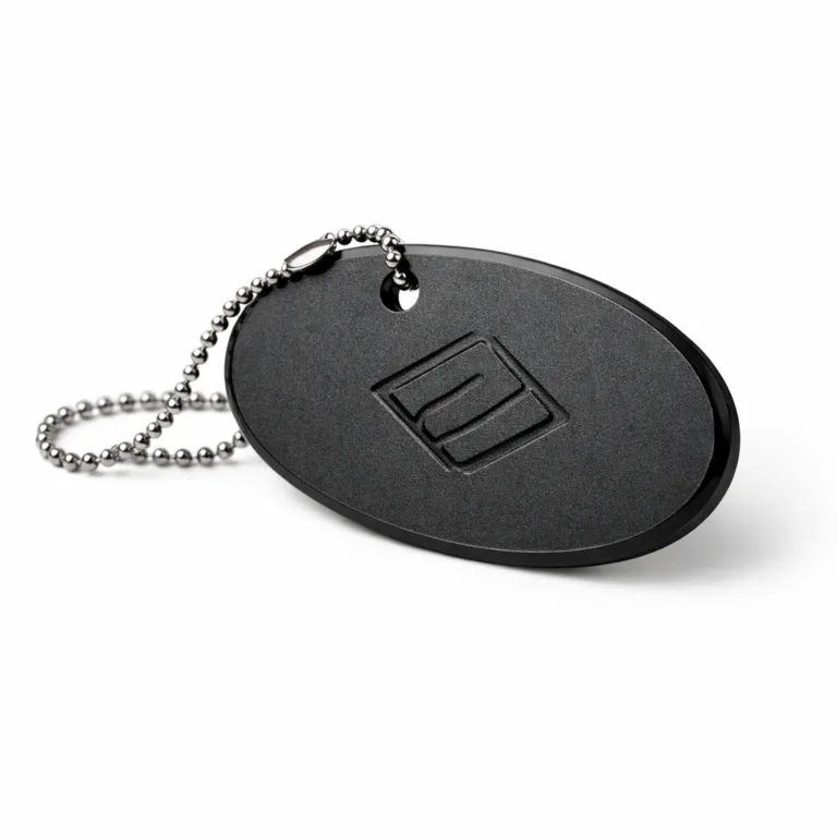 Oval luggage tag manufactured from matte black anodized aluminum, with sandblasted surface, debossed emblem, subtle chamfered edge, and attached with a ball chain.