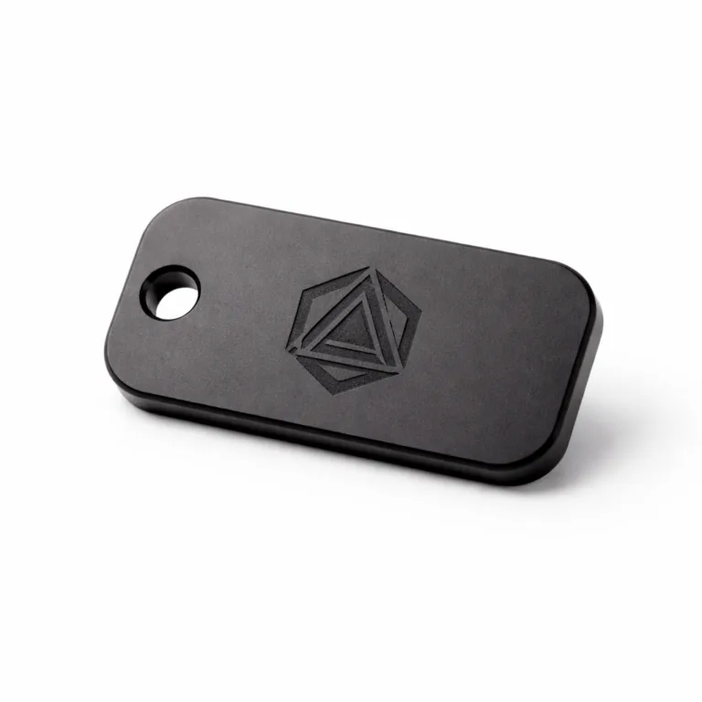 A realistic photograph of a matte black anodized aluminum logo tag for serial marking, with rounded corners, one mounting hole, and a precise laser-engraved logo shape.