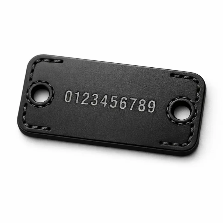 A matte black anodized aluminum garment tag, rectangular with rounded corners, two sewing holes, and etched details as well as stamping. Made for durable and stylish serial number marking on garments.