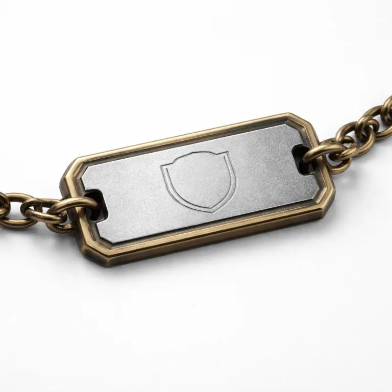A two-tone bracelet tag crafted from anodized aluminum and antique brass. It features a beveled edge, sandblasted matte texture, and a debossed blank emblem design, shot on a white background.