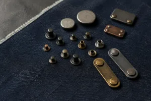A photo-realistic arrangement of custom rivets and metal tags on raw denim, showcasing the diverse finishes and applications for jeans.